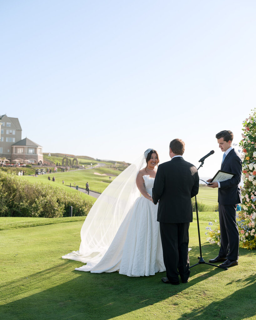 Bride and groom during their wedding ceremony at the Half Moon Bay Golf Links, with a view of the Ritz Carlton