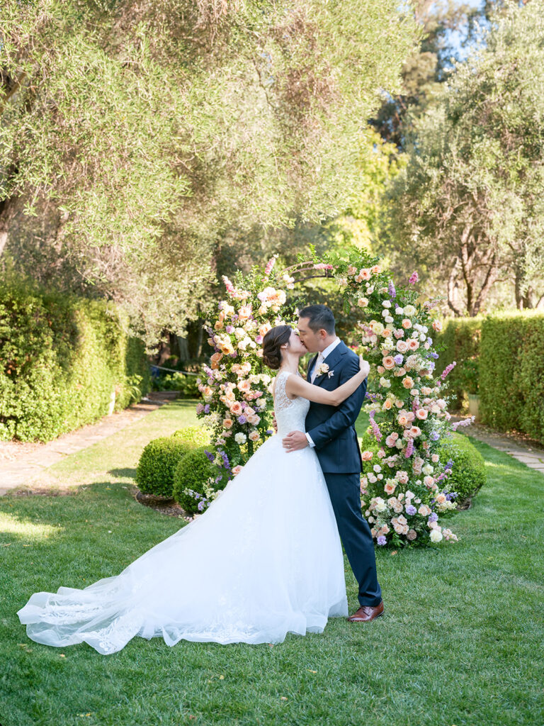 Bride and groom kiss in front of their floral wedding arch at the Allied Arts Guild, a Bay Area wedding venue in Menlo Park California