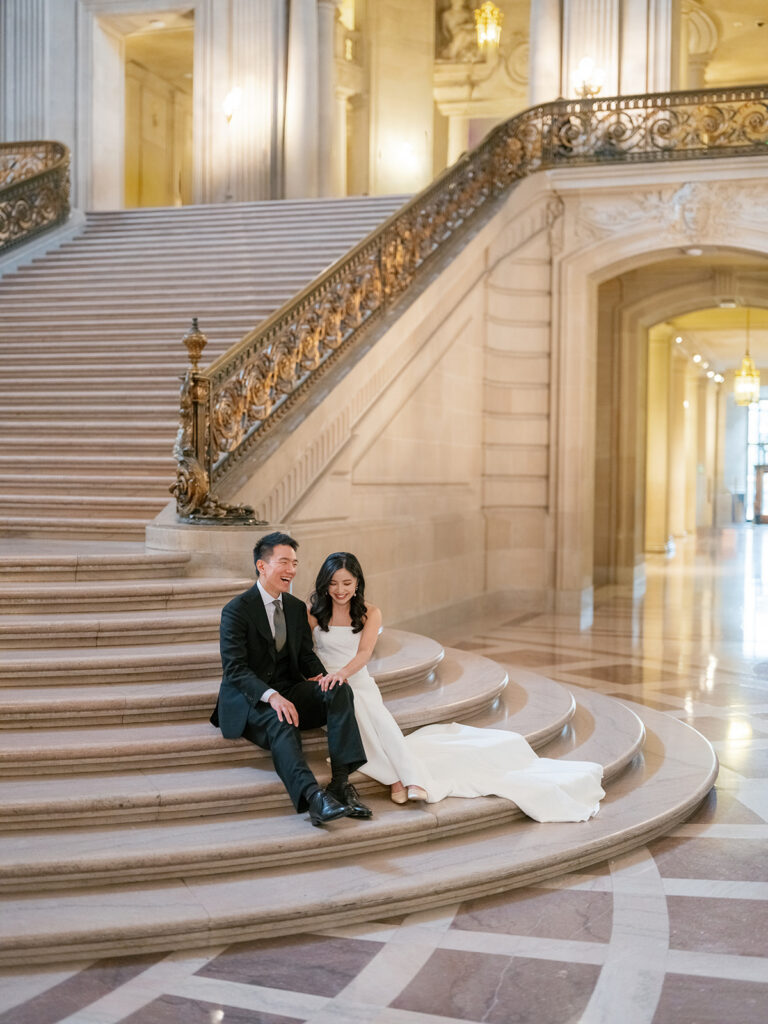 bride and groom sitting and laughing together at the grand staircase of San Francisco City Hall