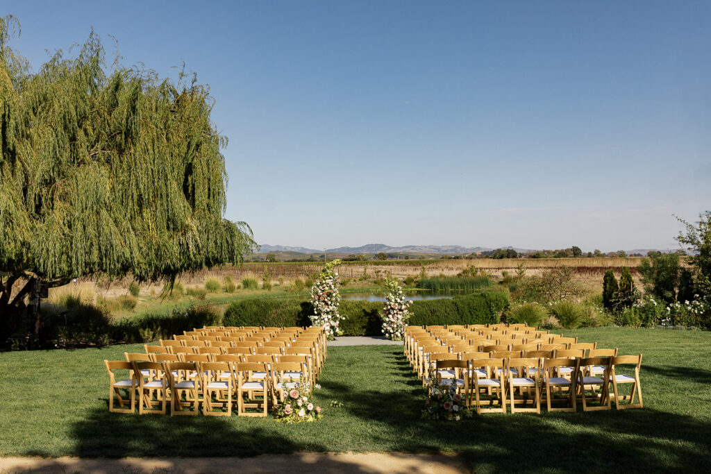 Outdoor ceremony setup at The Barn at Harrow Cellars — golden wooden chairs on green lawn, willow tree to the left, pond and Sonoma valley in the background