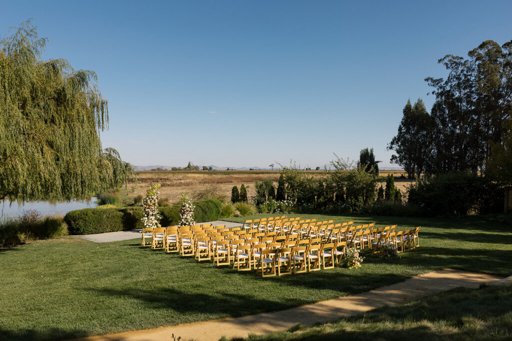 Angled view of the outdoor ceremony setup at The Barn at Harrow Cellars — golden wooden chairs on green lawn and floral arch in the background