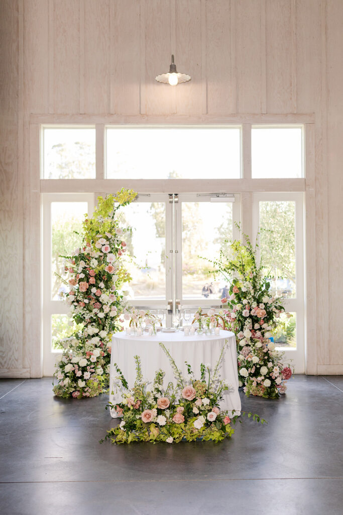 Sweetheart table inside The Barn at Harrow Cellars reception, flanked by tall floral columns of pink and white roses, floor-to-ceiling windows behind
