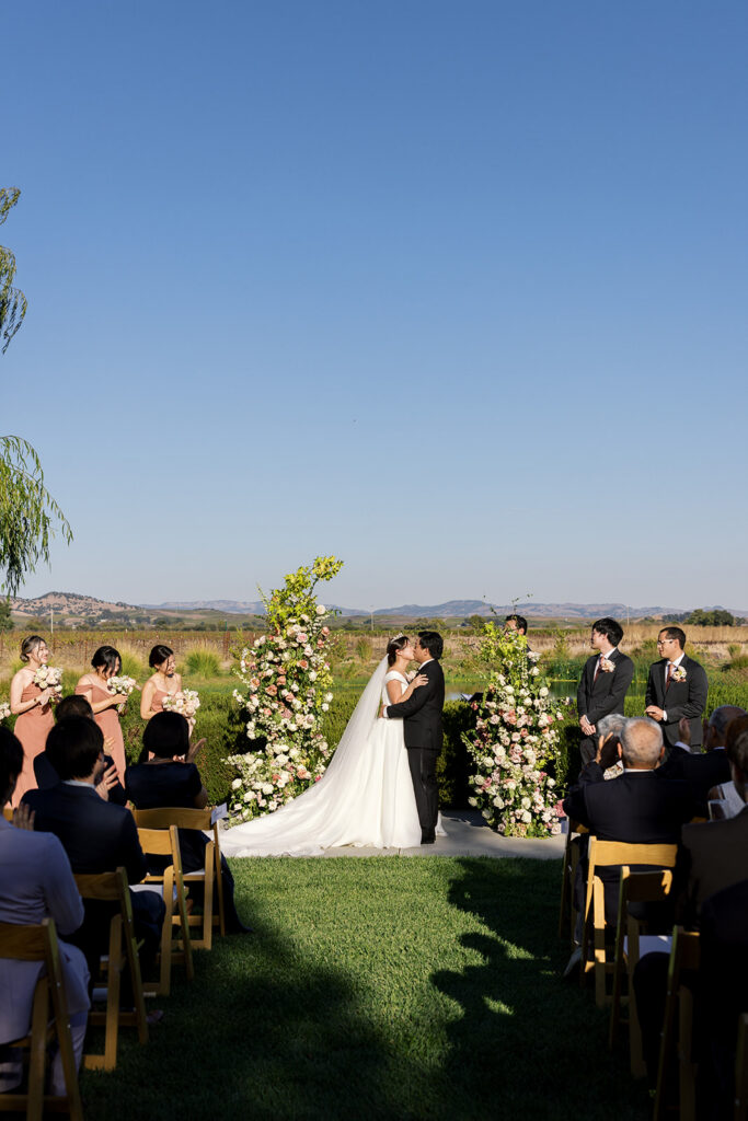 Bride and groom sharing their first kiss at The Barn at Harrow Cellars outdoor ceremony, Sonoma hills and vineyard stretching behind them — Sonoma wedding photographer