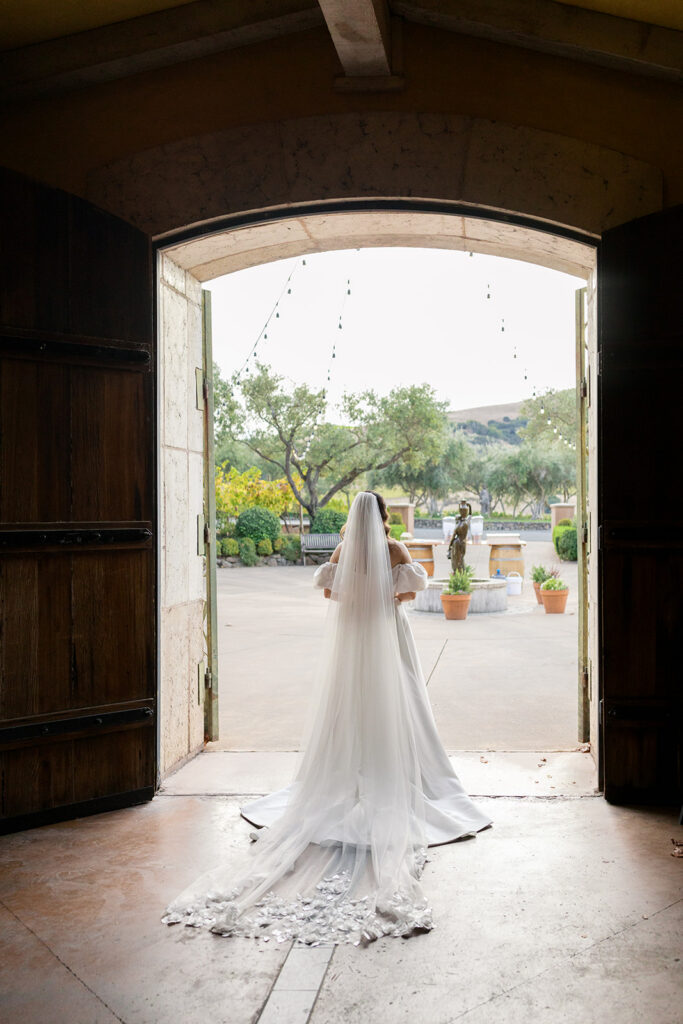 Bride in flowing white gown and cathedral veil standing in open wooden barn doors at Viansa Winery, looking out over the courtyard gardens — Sonoma wedding photographer