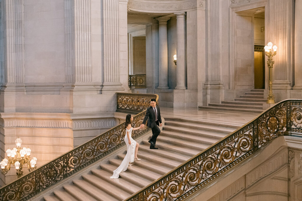 Bride and groom climbing the grand staircase hand in hand, ornate gold railing and warm sconce light at San Francisco City Hall