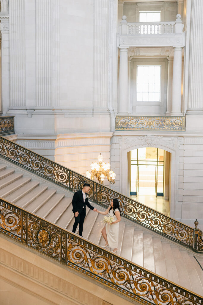 Bride and groom climbing the grand staircase hand in hand, ornate gold iron railing and arched ceiling at San Francisco City Hall