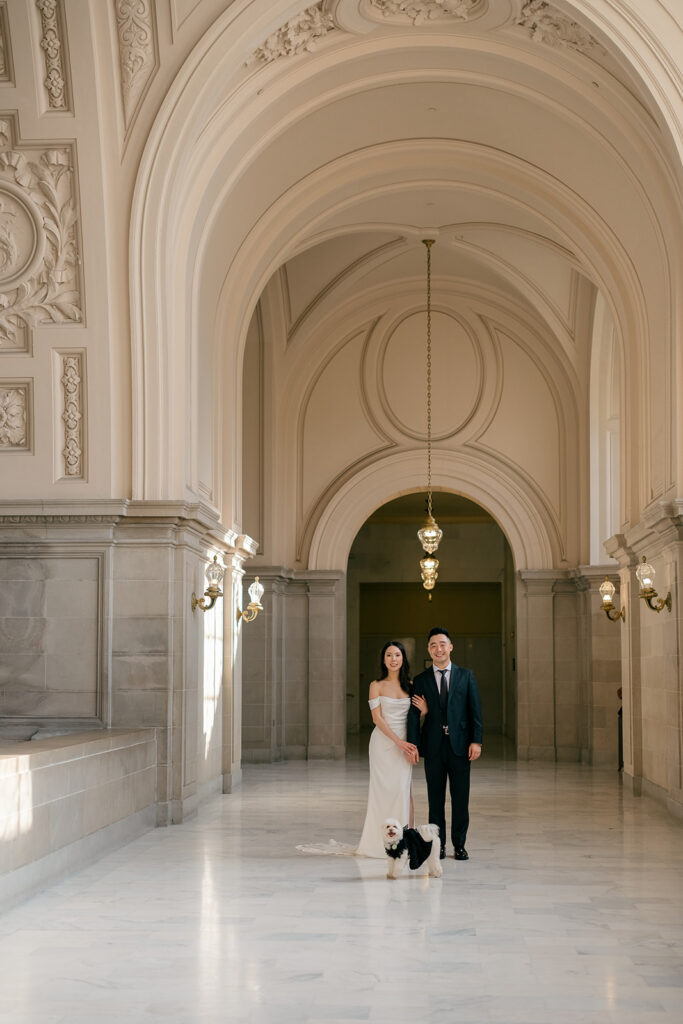 Bride and groom with their dog in the fourth floor hallway at San Francisco City Hall, arched ceiling and chandelier overhead