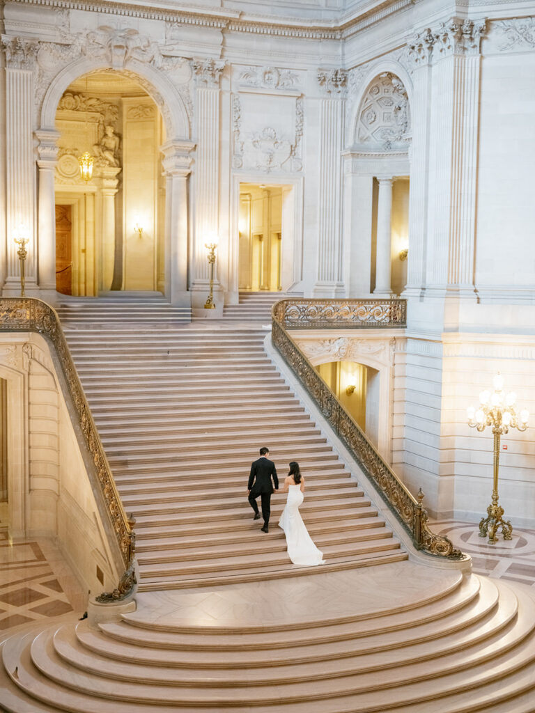 Wide aerial view of bride and groom ascending the grand staircase at San Francisco City Hall, gold iron railings flanking both sides