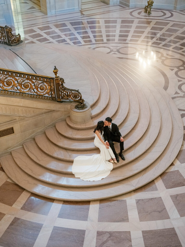 Overhead shot of bride and groom leaning in for a kiss on the curved base of the grand staircase, San Francisco City Hall wedding photography
