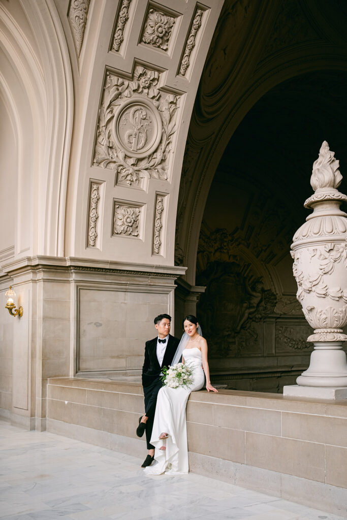 Groom in black suit and bride in white sleeveless dress holding a white bouquet beside the stone urn on the exterior colonnade of San Francisco City Hall