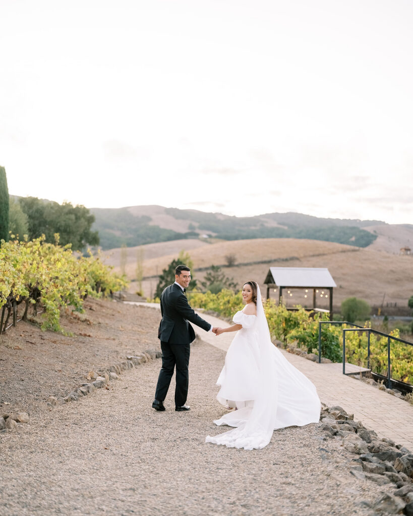 Bride and groom holding hands on a gravel vineyard path at Viansa Winery at golden hour, Sonoma rolling hills glowing behind them