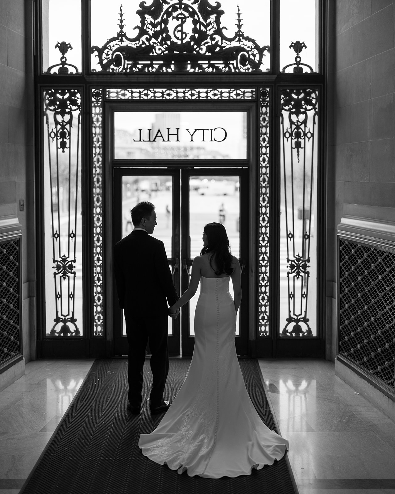 Black and white silhouette of bride and groom holding hands at the ornate iron City Hall entrance doors, San Francisco
