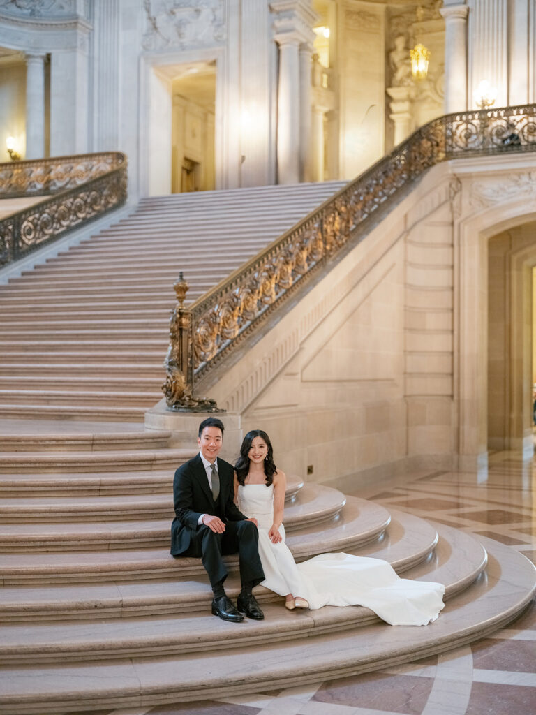 Bride and groom sitting together on the grand staircase steps at San Francisco City Hall, smiling at the camera