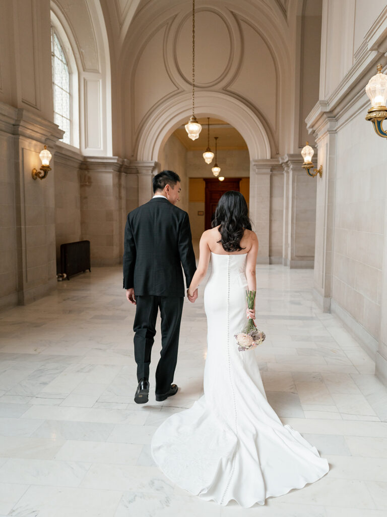 Bride and groom walking hand in hand down the fourth floor hallway at San Francisco City Hall, warm chandelier light overhead