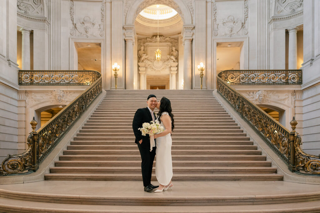 Wide shot of bride and groom small against the dramatic scale of the grand staircase at San Francisco City Hall