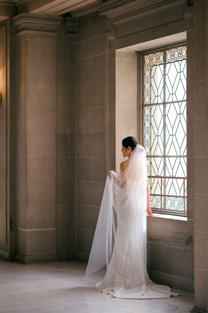 Bride with her back towards the camera next to a window with natural sunlight streaming in at San Francisco City Hall