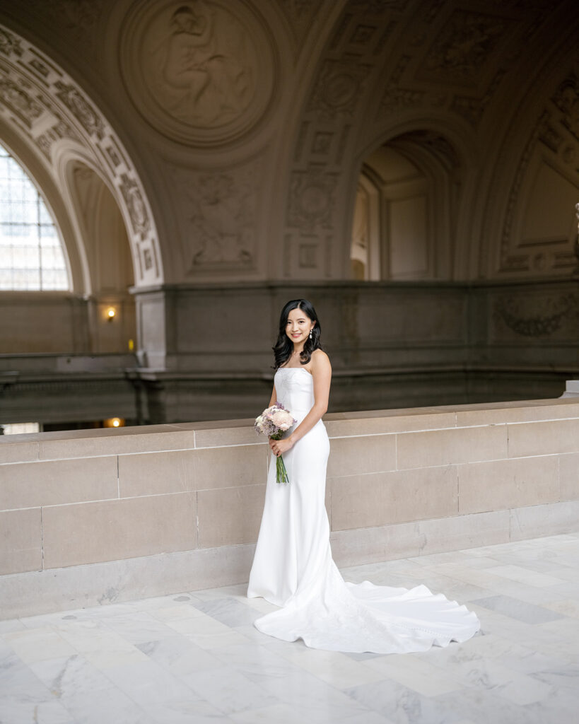Bride in strapless white gown with cathedral train holding a blush bouquet under the rotunda arch at San Francisco City Hall