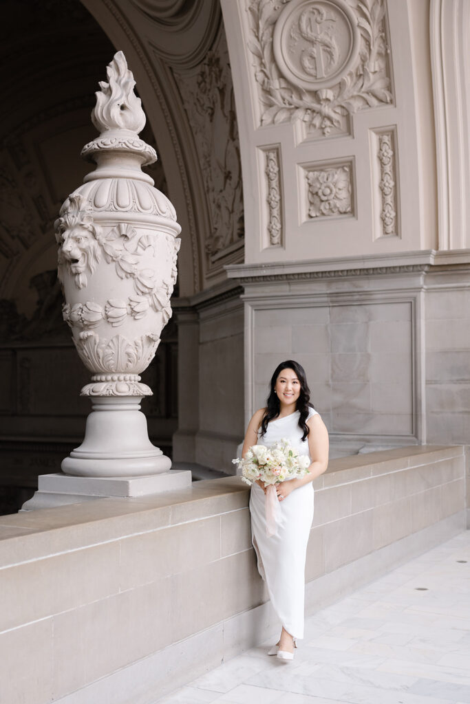 Bride in white sleeveless dress holding a white bouquet next to the ornate stone urn under the colonnade at San Francisco City Hall