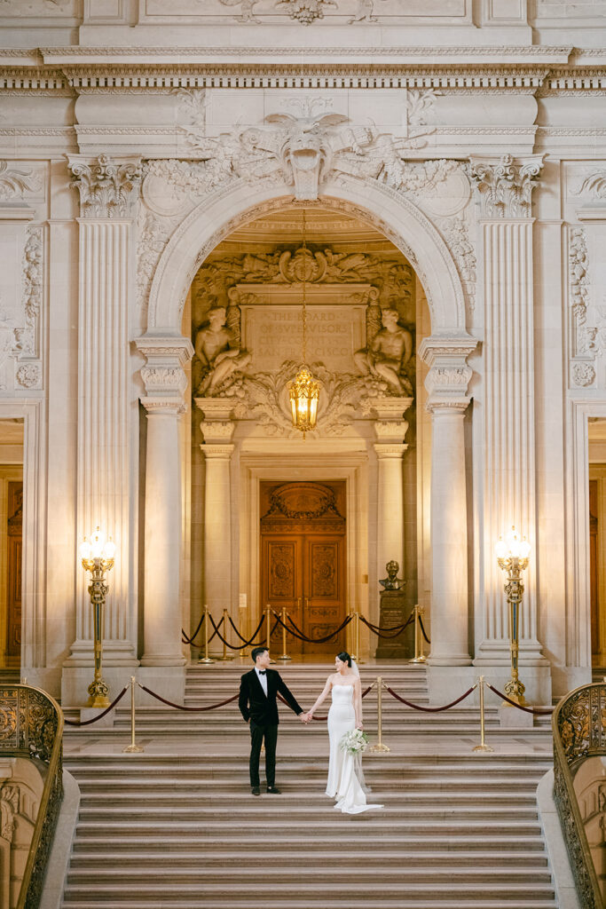 Bride and groom portrait at the base of the grand staircase, ornate arched ceiling and iron doors behind them, San Francisco City Hall wedding photographer
