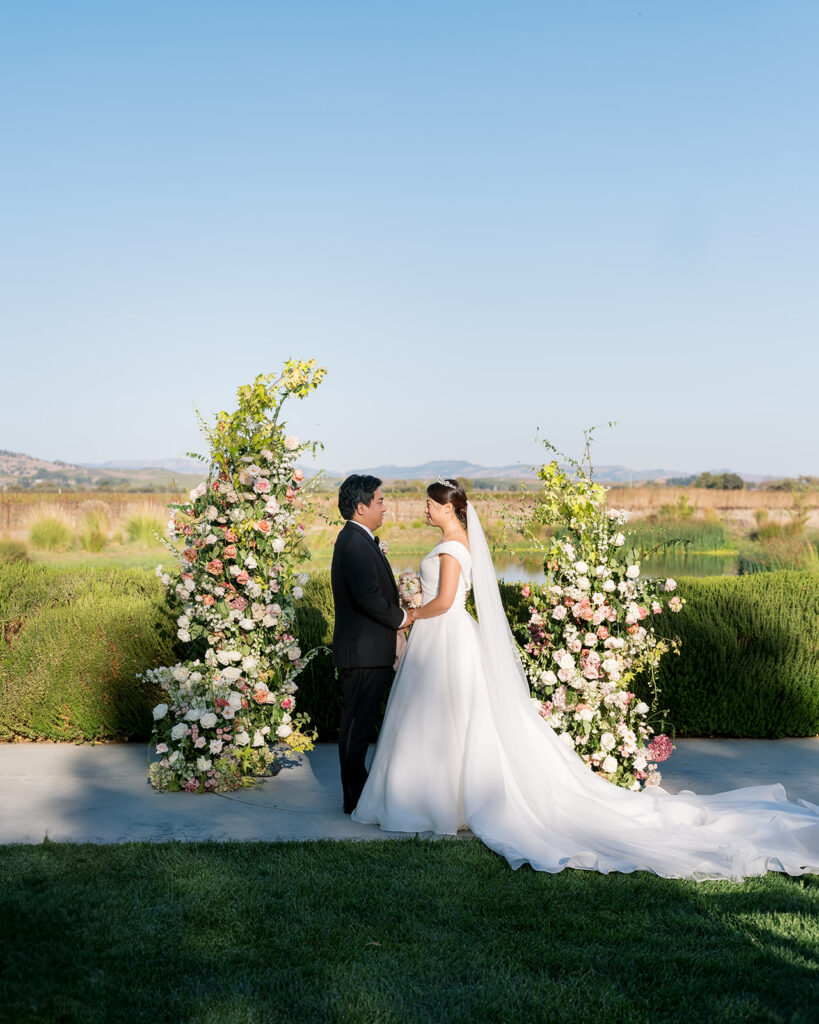 Bride and groom portrait at the floral arch at The Barn at Harrow Cellars, surrounded by pink and white roses with the pond and Sonoma hills behind them