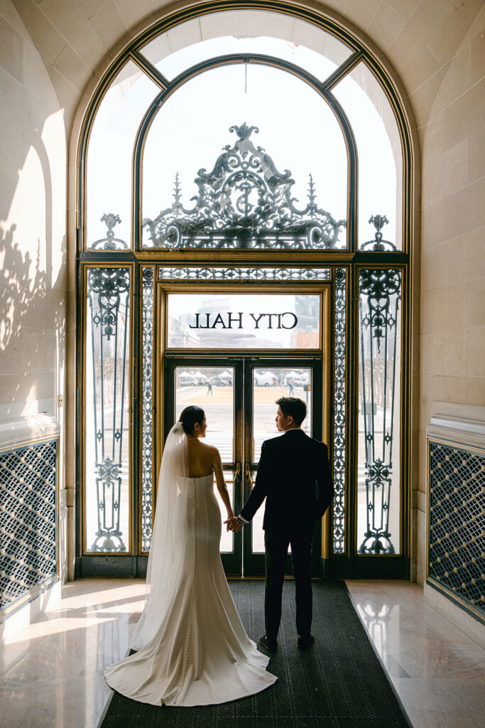 Bride and groom with backs turned towards the camera facing the ornate teal and gold main entrance doors of San Francisco City Hall