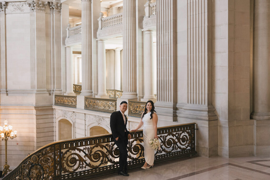 Bride and groom portrait on the upper staircase landing with tall Corinthian columns and gold railing at San Francisco City Hall