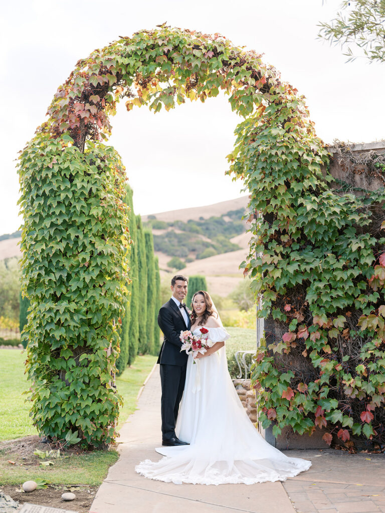 Bride and groom under a lush ivy and grapevine arch at Viansa Winery, Sonoma hills and cypress trees behind them — Sonoma wedding photography by sustudio