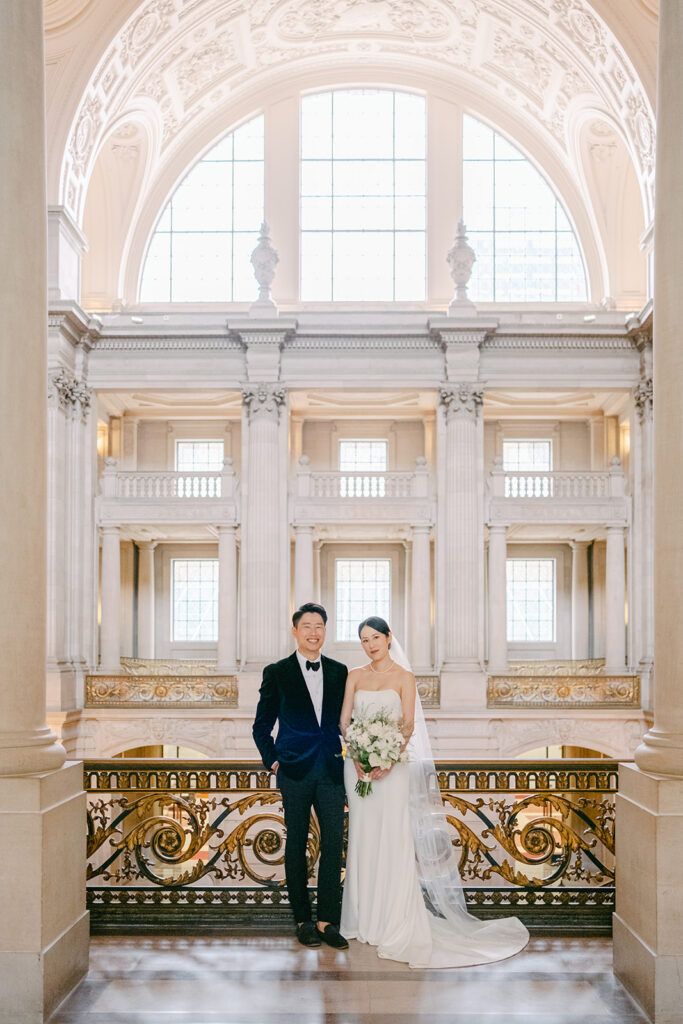 Bride and groom smiling on the upper grand staircase landing, tall columns and gold railing at San Francisco City Hall