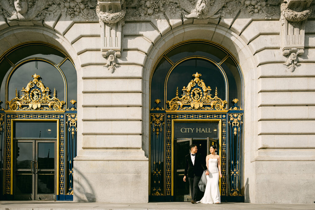 Bride and groom walking past the ornate teal and gold exterior doors of San Francisco City Hall after their wedding ceremony