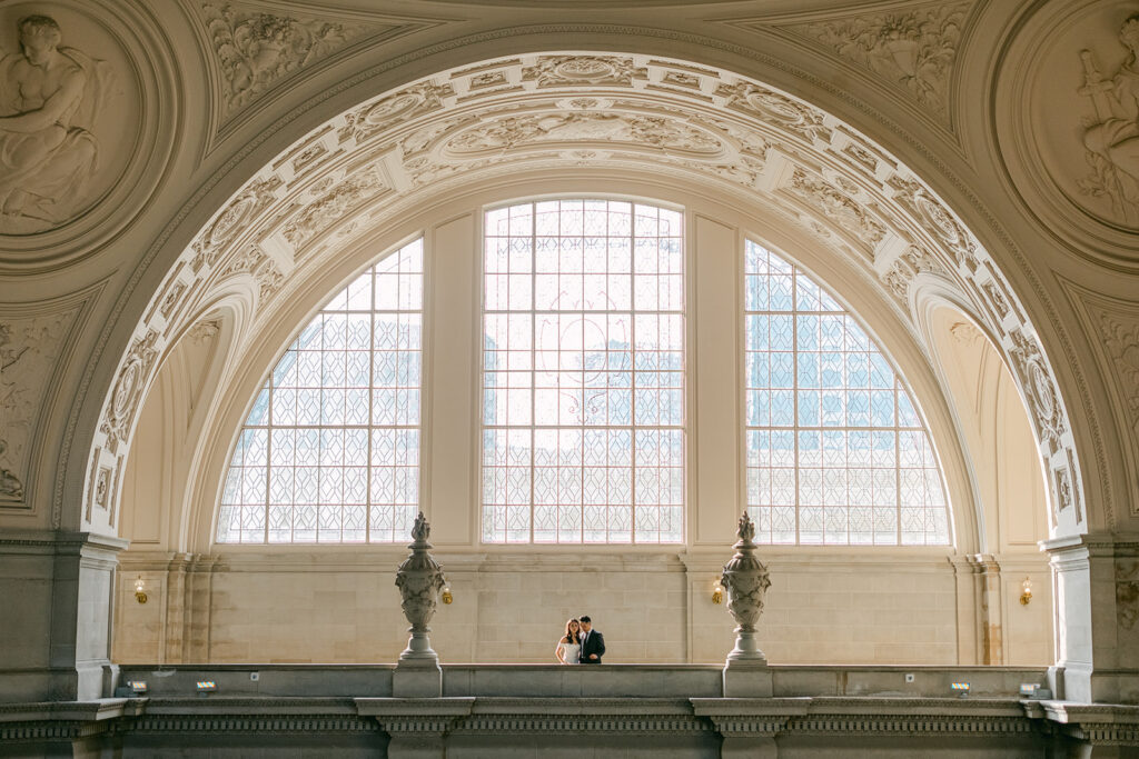 Wide shot of bride and groom standing small beneath the grand arched window on the fourth floor balcony at San Francisco City Hall — San Francisco City Hall wedding photography