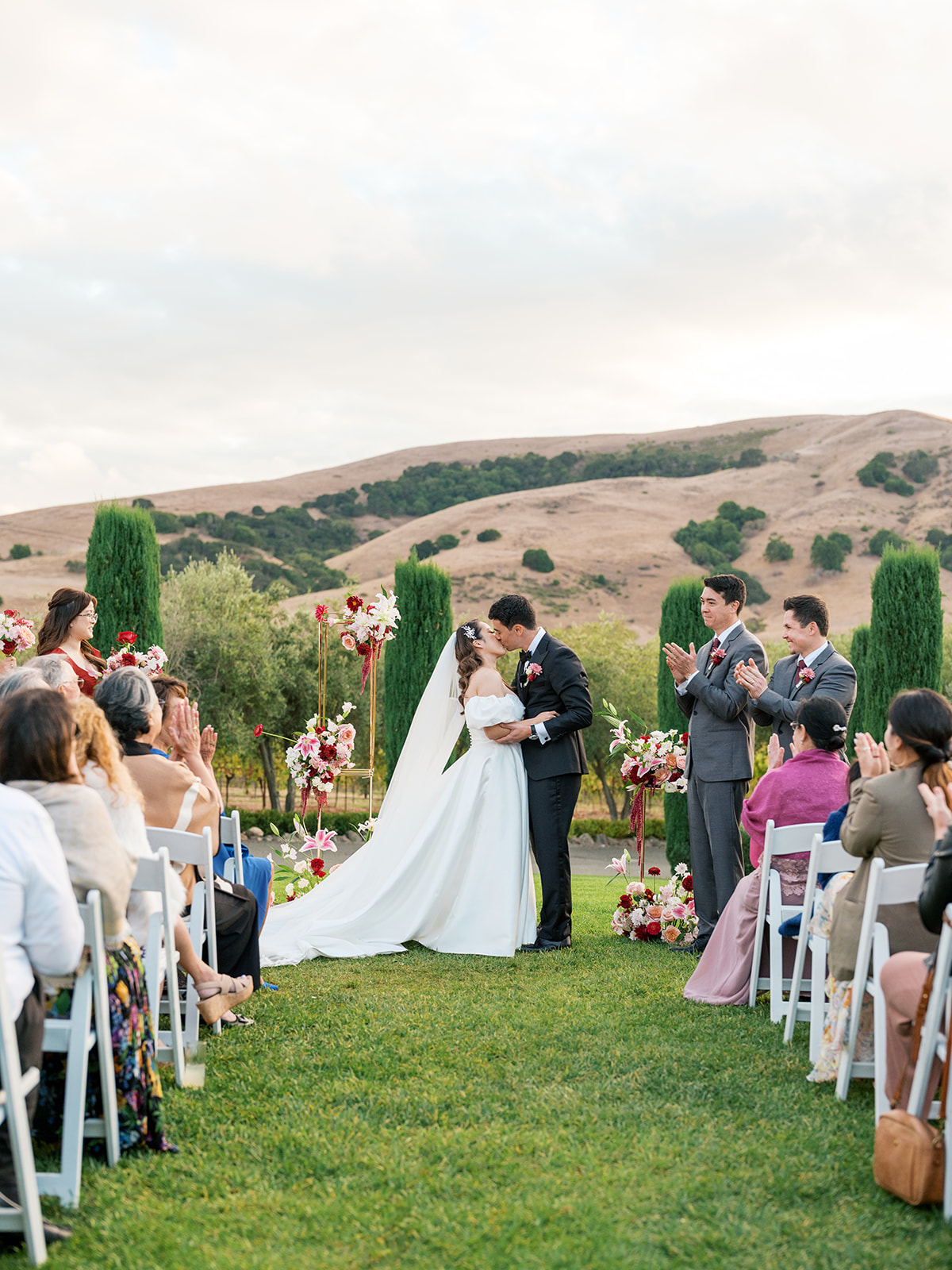 Bride and groom sharing their first kiss at Viansa Winery, guests applauding, Sonoma golden hills stretching behind them — Sonoma wedding photographer