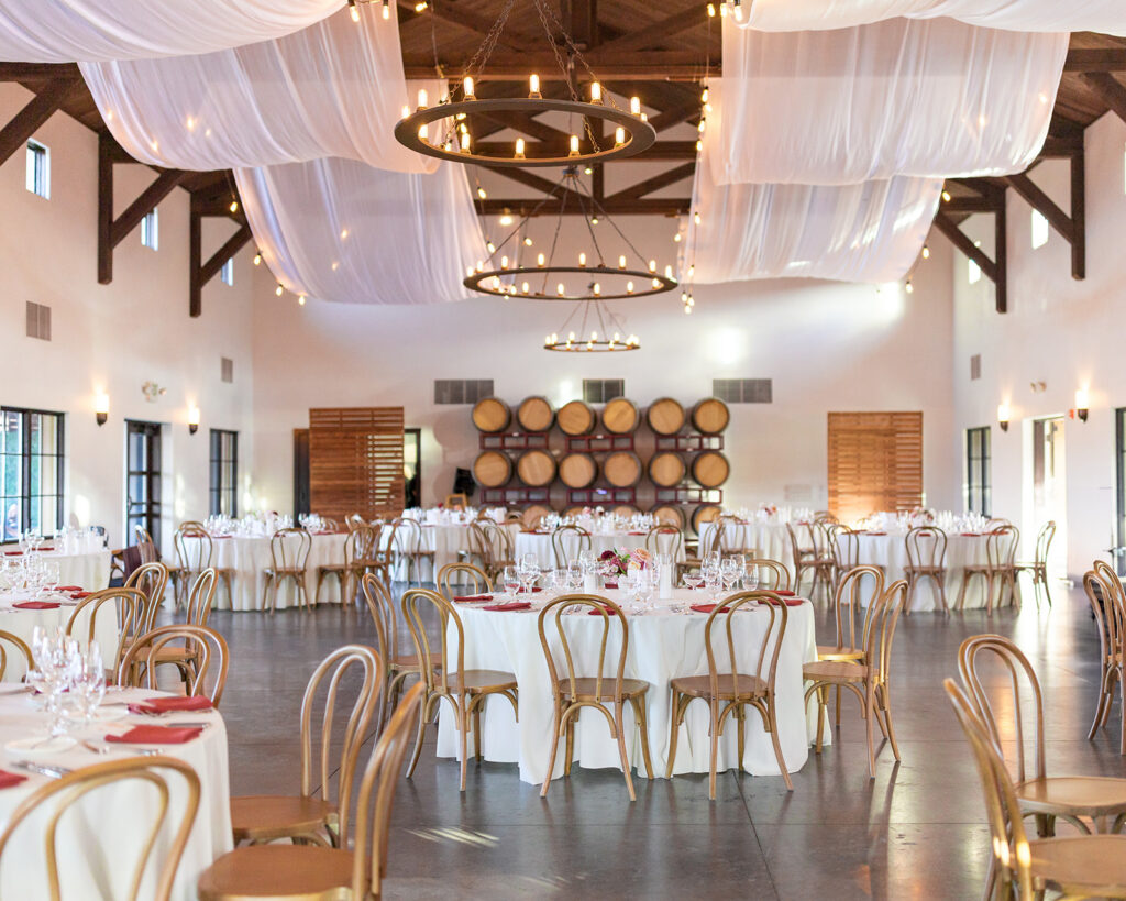 Viansa Winery reception hall set for a Sonoma wedding dinner — white draped ceiling, round tables with red napkins, gold crossback chairs, and a wine barrel wall backdrop