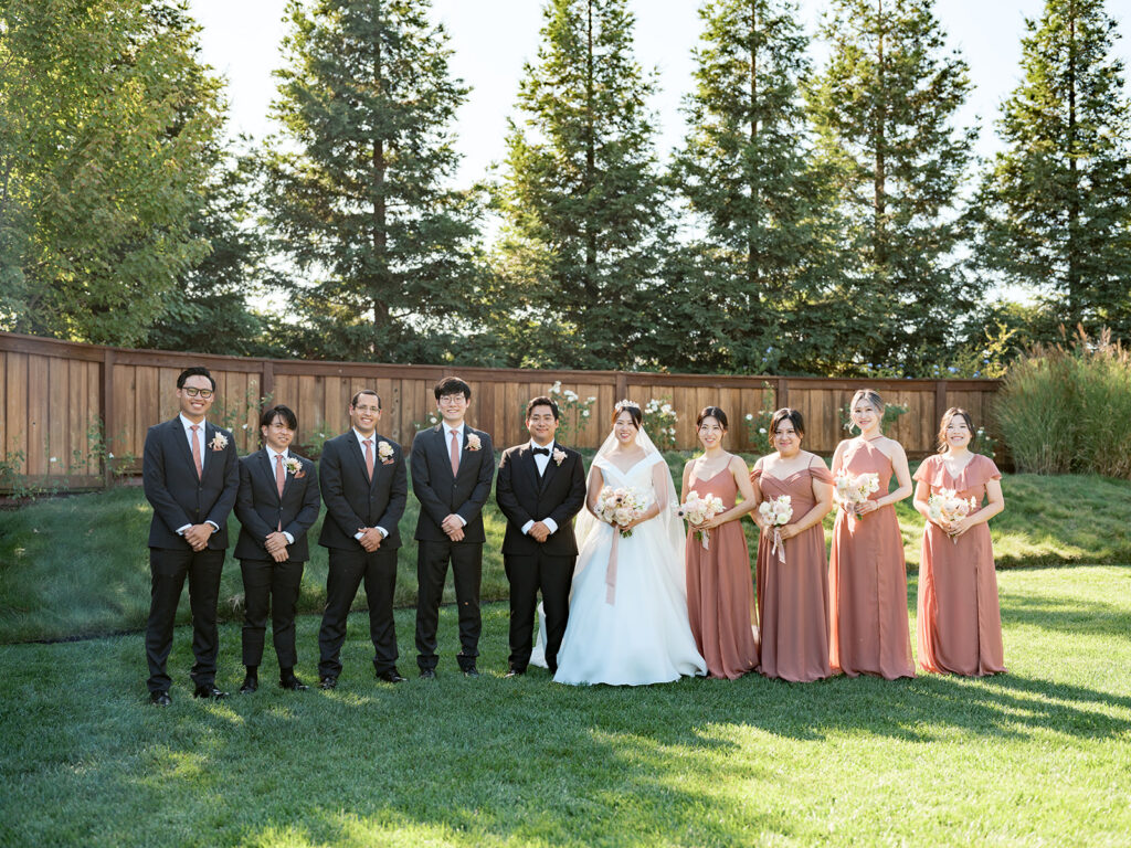 Wedding party portrait on the lawn at The Barn at Harrow Cellars in Sonoma — groomsmen in dark suits, bridesmaids in dusty rose, pine trees behind