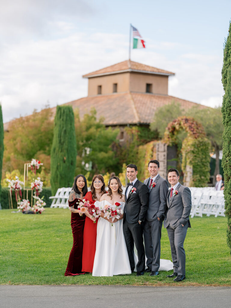Wedding party portrait at Viansa Winery in Sonoma, bridesmaids in deep red dresses and groomsmen in grey suits flanking the bride and groom on the lawn
