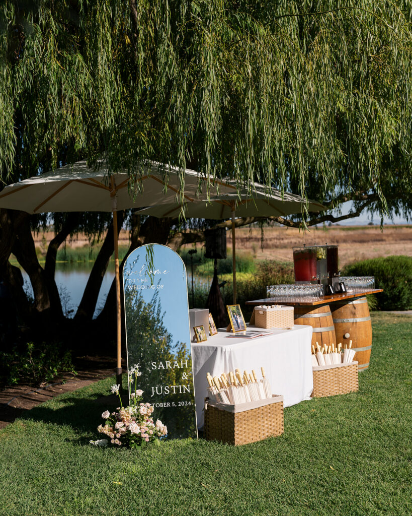 Welcome sign and bar setup under a willow tree at The Barn at Harrow Cellars Sonoma wedding, wine barrel bar and pond view behind